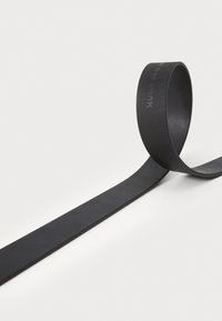 Bracelet noir en caoutchouc, plat et flexible, avec une texture lisse et un bord arrondi. Branding minimal embossé sur toute la longueur.
