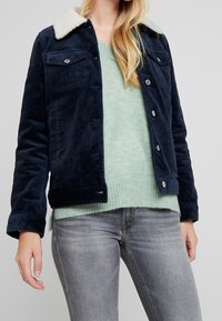 Marinblå kordjacka med faux shearling-krage, med knappstängningar och bröstfickor, bärs över en ljusgrön tröja och grå jeans.