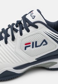 Weißer Sportschuh mit marineblauen Akzenten, ausgestattet mit einem strukturierten Mesh-Obermaterial, gerilltem Design und einem auffälligen Logo an der Seite.