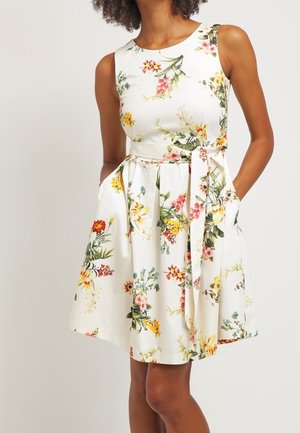 Vestido floral con fondo blanco, diseño sin mangas, cintura ajustada y bolsillos. Presenta coloridos patrones florales y un cinturón de lazo.