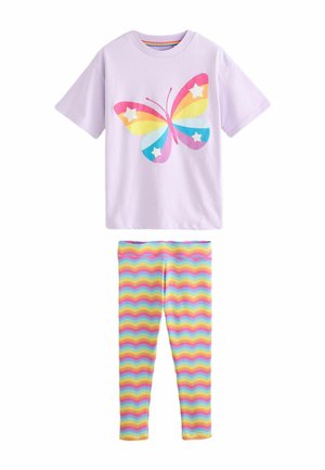 Glimlachend meisje met lang donker haar draagt een lavendelkleurig shirt met kleurrijke vlinderprint en legging met regenboog zigzag patroon en witte sneakers.