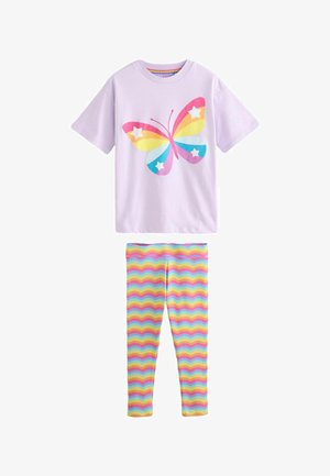 Glimlachend meisje met lang donker haar draagt een lavendelkleurig shirt met kleurrijke vlinderprint en legging met regenboog zigzag patroon en witte sneakers.