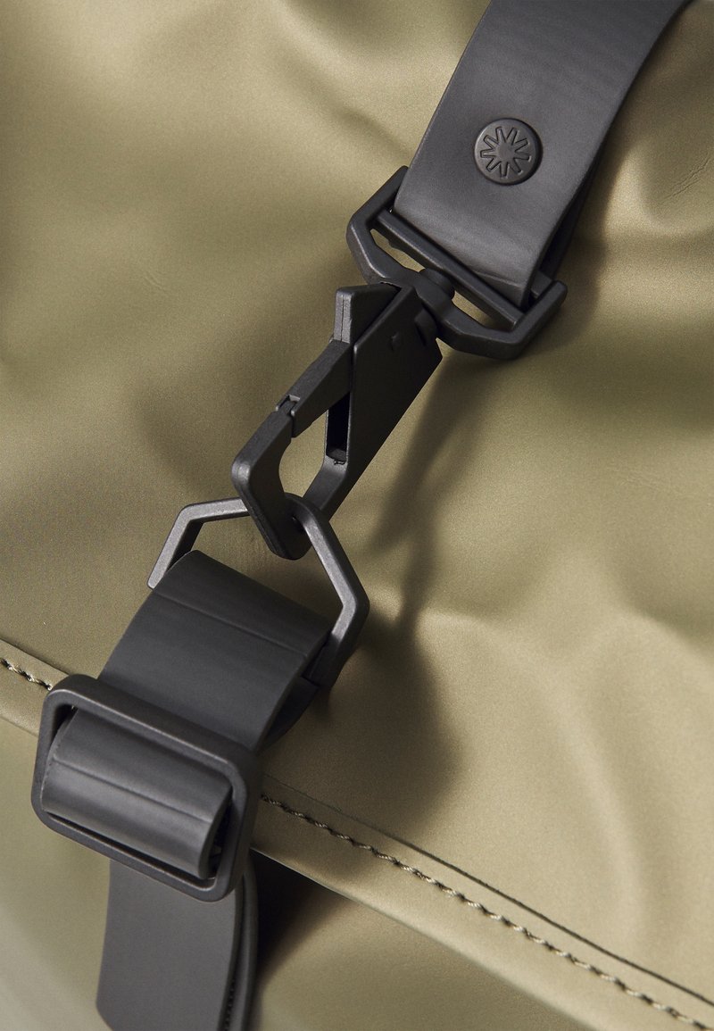 Clip et sangle en plastique noir sur un tissu olive lisse. Comprend une boucle noire texturée et un emblème de marque identifiable.