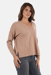 Maglione a V-collo beige, in maglia, con maniche lunghe, caratterizzato da una vestibilità comoda e spacchi laterali. Texture morbida, design casual abbinato a jeans scuri.