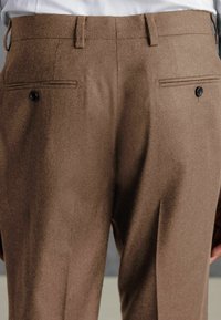 Pantalones marrones a medida con una textura suave, dos bolsillos en la parte posterior y detalles en botones negros. Presentan un frente plano y una forma estilizada.