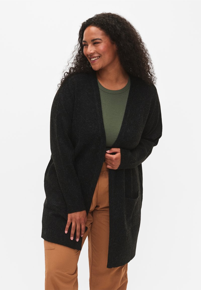 Schwarze Strickjacke mit offenem Front, langen Ärmeln und seitlichen Taschen, getragen über einem grünen Oberteil und kombiniert mit beigefarbenen Hosen. Strukturiert.