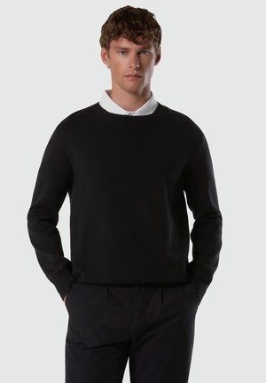 Mann trägt schwarzen Pullover mit rundem Ausschnitt über weißem Hemd mit Kragen, schwarze Hose, steht mit Händen in den Taschen vor hellem grauem Hintergrund.
