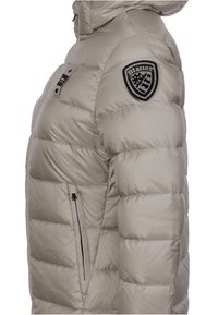 Beige gesteppte Jacke mit einer Reißverschlusstasche, gesteppter Verzierung und einem schwarzen Aufnäher mit weißem Design auf der linken Schulter. Gepolstert für Wärme.