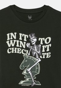 Juodas medvilnės marškinėliai su grafika, kurioje pavaizduotas skeletas su cilindru, laikantis kortas, šachmatų figūra ir tekstas "IN IT TO WIN IT" kontrastingomis spalvomis.