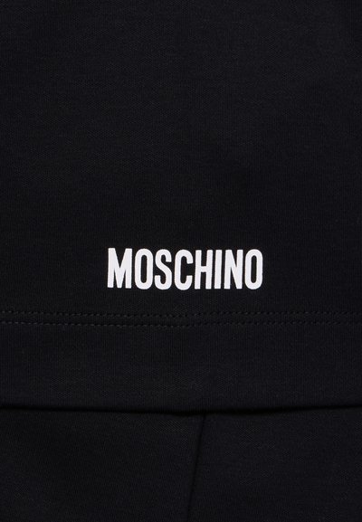 Melnas auduma pamatne ar horizontālu baltu teksta logotipu "MOSCHINO" apakšējā daļā; gluda tekstūra ar redzamām šuvju detaļām.