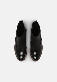 Botas negras hasta el tobillo que presentan una combinación de texturas mate y brillantes, con perforaciones decorativas y un diseño en forma de ondas a lo largo de la parte superior.