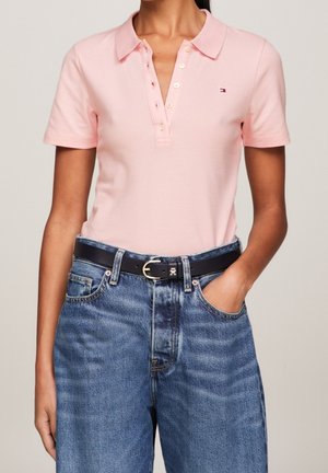 Donna che indossa una polo rosa chiaro a maniche corte infilata in jeans blu a vita alta con cintura nera, mano in tasca.