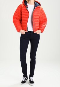 Chaqueta acolchada roja con capucha, con un forro azul marino y cierre de cremallera, combinada con jeans oscuros y zapatillas negras. Textura suave, diseño acolchado.