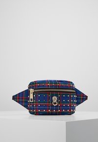 Blå och röd rutmönstrad tygväska med nitar, guldzipper och ett dekorativt spänne. Har en platt botten och justerbar rem.