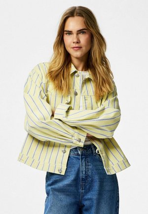 Femeie cu păr lung blond, purtând o jachetă cu dungi galbene și albe și blugi albaștri, stând cu brațele încrucișate în fața unui fundal alb.