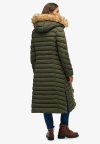 Lange, olijfgroene pufferjas met een nepbont capuchon, voorzien van horizontale stiksels, zijsplitsingen en gecombineerd met bruine veterschoenen.
