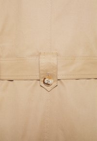 Beige stof met een gladde textuur, voorzien van een brede riem met een spitse lip en een ronde knoop met vier gaten als sluitdetail.