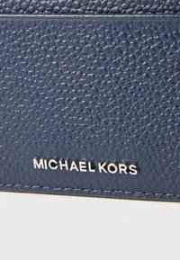 Laivastonsininen teksturoitu nahka, jossa on sileä pinta ja hienovarainen kuvio. Alareunassa on hopeanvärinen "MICHAEL KORS" -logo painettuna.