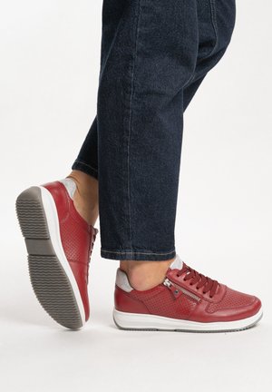 Person wearing dark jeans and red leather sneakers with white soles and side zippers, walking on a white surface.