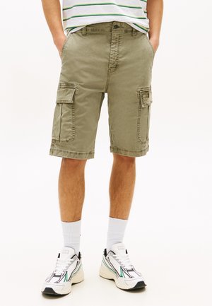 Mand iført olivengrønne cargoshorts, hvide sokker og hvide sneakers, stående med hænderne i lommerne.