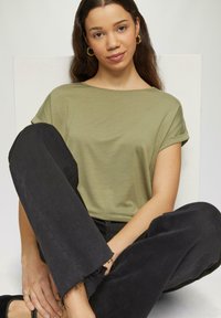 Oliwkowa koszula z krótkim rękawem i podwiniętymi mankietami, w połączeniu z czarnymi spodniami dżinsowymi o szerokich nogawkach z postrzepionymi brzegami. Model siedzi na tle w jasnych kolorach.