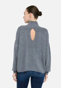 Vrouw met een knot in haar haar, gekleed in een grijze geribde trui met een keyhole-detail aan de achterkant en blauwe spijkerbroeken, met haar rug naar de camera gekeerd tegen een witte achtergrond.