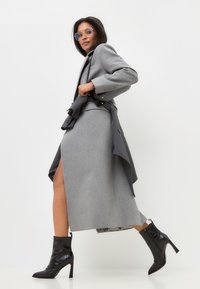 Cappotto in lana grigio con uno spacco alto e design su misura, abbinato a stivaletti neri con tacco. Caratterizzato da una silhouette aderente e spalle strutturate.