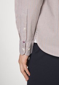 Gros plan sur le bras d'une personne portant une chemise beige et blanche rayée avec des poignets boutonnés, à côté d'un pantalon foncé sur un fond uni.