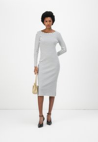 Robe ajustée à manches longues en blanc avec des rayures noires ; décolleté rond, longueur genou, accompagnée d'un sac à main beige et de talons noirs.