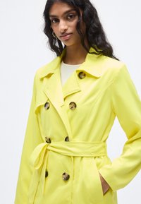 Φωτεινό κίτρινο παλτό από λείο ύφασμα, με διπλό κούμπωμα, μεγάλα κουμπιά και ζώνη στη μέση που δένει για σιλουέτα εφαρμοστή.