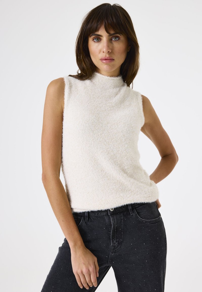 Jonge vrouw met bruin haar, gekleed in een mouwloos wit topje met structuur en een zwarte high-waisted spijkerbroek met subtiele witte spikkels.