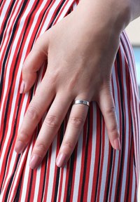 Silberner Ring an einer Hand mit heller Haut. Die Hand liegt auf einem Stoff mit roten und weißen vertikalen Streifen und schwarzen Akzenten. Die Nägel sind in einem blassen Rosa lackiert.