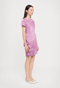 Robe satinée violette avec des manches courtes, détail froncé sur le côté et liens ajustables. Associée à des mules blanches à talons.