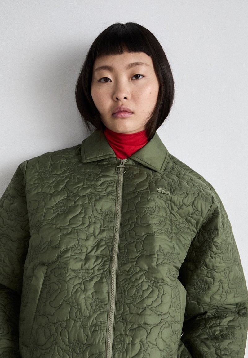 Grüne Steppjacke mit floralem Stitching, Frontreißverschluss und spitzem Kragen, getragen über einem roten Rollkragenpullover. Glatte Textur.