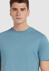 T-shirt azzurro chiaro a maniche corte realizzato in tessuto morbido, con scollatura rotonda e un piccolo logo arancione sul lato sinistro del petto.
