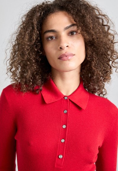 Röd ribbad tröja med krage, försedd med sex silverfärgade knappar framtill. Mjuk textur och figurnära design.