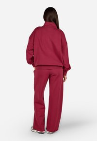 Vīna sarkanā lielā izmēra sporta krekls un atbilstoši platās bikses, izgatavotas no mīksta auduma ar ribotām aprocēm un jostu. Apavi redzami apakšā.