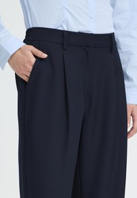 Pantalon bleu marine avec une texture lisse, présentant des plis à l'avant, des poches latérales et une ceinture ajustée. Le tissu semble léger et élégant.