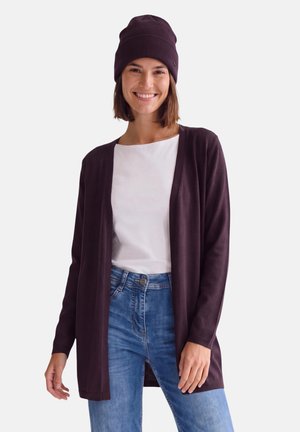 Lächelnde Frau mit dunkelviolettem Beanie, passendem offenem Cardigan, weißem Hemd und blauer Jeans vor einem einfarbigen hellen Hintergrund.