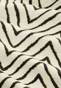 Tissu crème avec des rayures en zigzag noires et dorées, présentant une surface texturée et un tombé souple, mettant en valeur le contraste audacieux du motif.