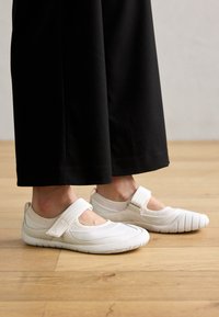 Scarpe slip-on bianche flessibili con una fascetta, dotate di una tomaia liscia, dettagli testurizzati sulla punta e una suola in gomma, indossate con pantaloni neri.