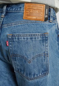 Jeans in denim azzurro chiaro con etichetta in pelle che mostra il nome del marchio e il numero di lotto, caratterizzati da una tasca posteriore con etichetta rossa e cucitura a contrasto.