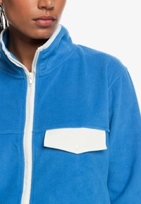 Blue fleece jacket with a high collar, white zip, and a white snap-button chest pocket. Soft texture, casual design.