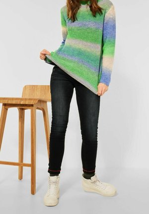 Person, der den Saum eines grün, blau und beige gestreiften Pullovers herunterzieht, trägt schwarze Jeans und weiße Sneakers und steht nahe einem Holzstuhl.