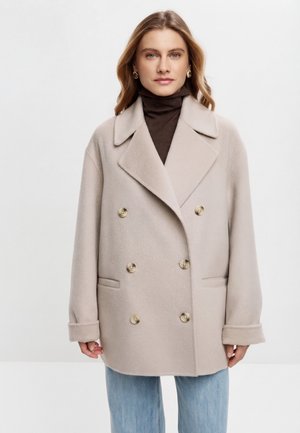 Kobieta o jasnobrązowych włosach ubrana w beżowy, dwurzędowy płaszcz na ciemnym golfie i jasnoniebieskich dżinsach, stoi na tle jednolitego białego tła.