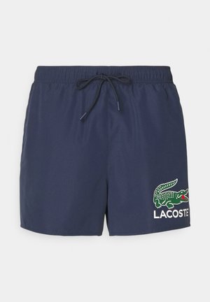 Tumedad sinises ujumispüksid, mis on valmistatud kergest kangast. Neil on elastne vöökoht, millel on pael ning roheline krokodilli logo koos "LACOSTE" tekstiga.