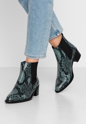Bottes à talons montant à la cheville avec un motif peau de serpent vert et noir, portées avec un jean court bleu clair sur un sol blanc.