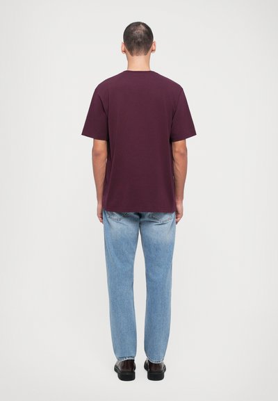 Camiseta de algodón color burdeos con mangas cortas, corte estándar y dobladillo recto, combinada con jeans de mezclilla azul claro y zapatos negros.