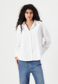 Valkoinen napillinen paita, jossa on rento leikkaus, pitkät hihat ja klassinen kaulus. Kangas näyttää kevyeltä ja teksturoidulta. Yhdistetty sinisiin farkkuihin.