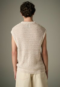 Un haut sans manches en tricot beige clair avec un motif de diamant texturé, un ourlet côtelé et une trame ouverte, associé à un pantalon large de couleur crème.